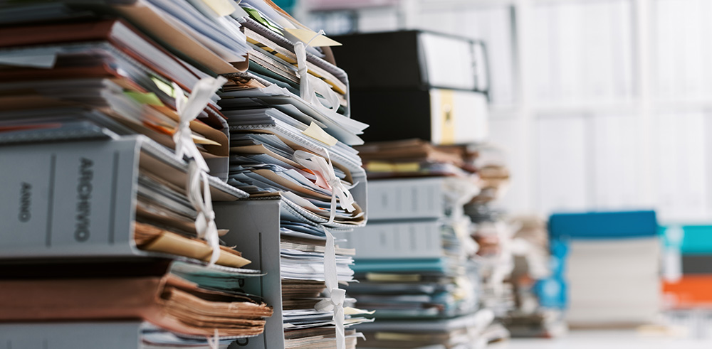 Scrivania colma di documenti e fascicoli impilati, con cartelle d'archivio etichettate in primo piano. Sullo sfondo, un ufficio luminoso con scaffali e faldoni colorati, rappresentando un ambiente di gestione documentale e archiviazione.