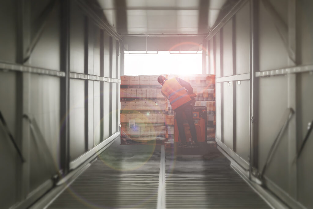 Operatore in magazzino che movimenta pallet all'interno di un camion, visto attraverso una grande lettera 'X' stilizzata, con effetti di luce e sfumature verdi. L'immagine rappresenta logistica, efficienza e il brand XTransport